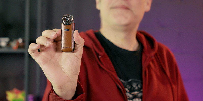 Eine Person in einem roten Hoodie hält ein kleines, elegantes Vape-Pod-System mit braunem Leder-Design in der Hand. Das Gerät wird in den Vordergrund gerückt, während das Gesicht der Person nur teilweise und unscharf zu sehen ist. Der Hintergrund ist dunkel und leicht verschwommen mit Regalen, auf denen verschiedene Gegenstände stehen.