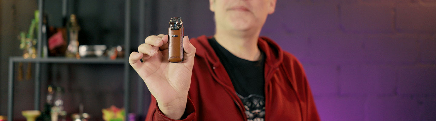 Eine Person in einem roten Hoodie hält ein kleines, elegantes Vape-Pod-System mit braunem Leder-Design in der Hand. Das Gerät wird in den Vordergrund gerückt, während das Gesicht der Person nur teilweise und unscharf zu sehen ist. Der Hintergrund ist dunkel und leicht verschwommen mit Regalen, auf denen verschiedene Gegenstände stehen.