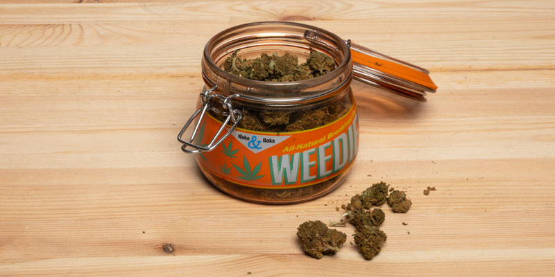 Ein ge&ouml;ffnetes Einmachglas mit der Aufschrift &laquo;WEED&raquo; auf einem Holzuntergrund. Das Glas ist mit getrockneten Cannabisbl&uuml;ten gef&uuml;llt, einige davon liegen daneben auf dem Tisch.