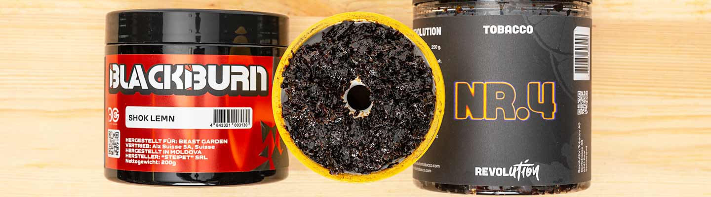 Zwei Dosen Shisha-Tabak der Marken Blackburn (Shok Lemn) und Revolution (NR.4) stehen nebeneinander, in der Mitte befindet sich ein gelber Shisha-Kopf mit befülltem Tabak in dunkler Farbe und Loch in der Mitte.