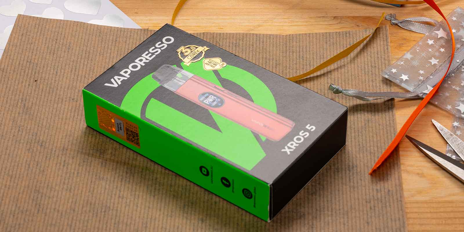 Vaporesso XROS 5 Pod-System in Originalverpackung auf Holzoberflaeche, Geschenkband und Schere im Hintergrund