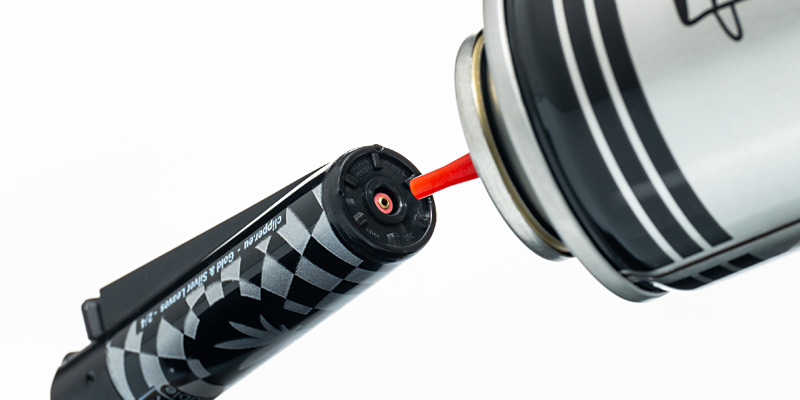 Ein Clipper-Feuerzeug mit schwarz-weissem Schachbrettmuster wird mit Gas nachgefüllt. Eine Nachfüllkartusche mit einer roten Düse ist in die Nachfüllöffnung am Boden des Feuerzeugs eingeführt. Die Szene zeigt den einfachen und nachhaltigen Nachfüllmechanismus der Clipper-Feuerzeuge.