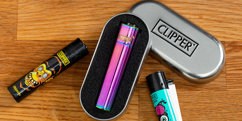 Eine Sammlung von Clipper-Feuerzeugen auf einer Holzoberfläche. In der Mitte befindet sich ein metallisch schimmerndes, buntes Clipper-Feuerzeug in einer silbernen Metallbox mit «CLIPPER»-Logo. Links daneben liegt ein schwarzes Feuerzeug mit einem auffälligen Totenkopfdesign, und rechts ein weiss-blaues Feuerzeug mit einem grafischen Muster. Die Szene betont die hochwertigen Designs und die Vielfalt der Clipper-Feuerzeuge.