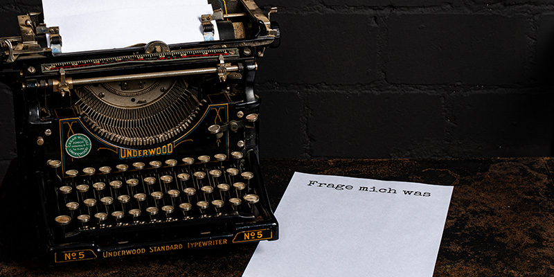 Alte Schreibmaschine auf schwarzer Matte mit weissem Papier mit Aufschrift &laquo;Frage mich was&raquo;.