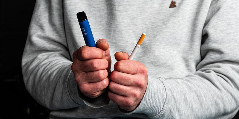 Person in grauem Pullover h&auml;lt in der linken Hand eine blaue Einweg-E-Zigarette und in der rechten Hand eine herk&ouml;mmliche Zigarette, mit angespannten F&auml;usten als Symbol f&uuml;r den Wechsel vom Rauchen zum Dampfen. Schwarzer Hintergrund.