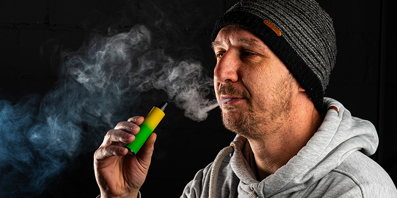 Mann mit grauem Hoodie und schwarzer Strickm&uuml;tze dampft eine Einweg-E-Zigarette mit gr&uuml;n-gelbem Design, w&auml;hrend er Rauch ausatmet. Schwarzer Hintergrund mit sichtbarem Dampfwolke-Effekt.