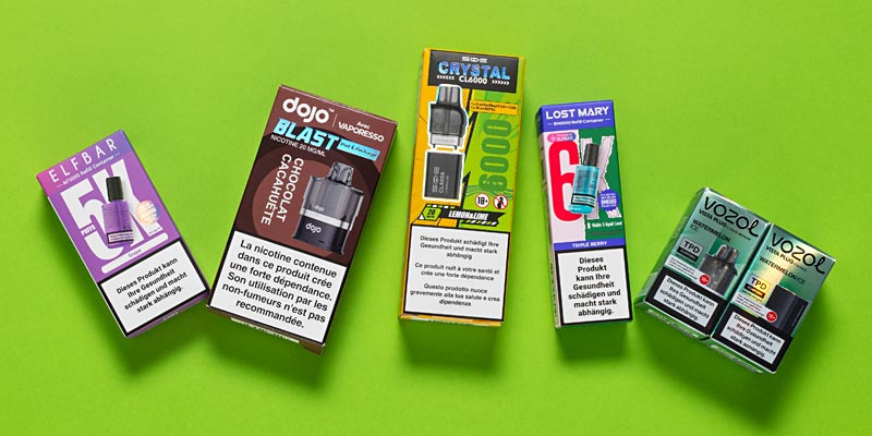 Eine Auswahl an verschiedenen Vape-Produkten liegt auf einem grünen Hintergrund. Von links nach rechts sind folgende Marken und Modelle sichtbar: Elfbar 1500 in Lila-Verpackung, Dojo Blast mit Schokoladen- und Erdnussgeschmack in einer braunen Verpackung, SKE Crystal CL000 in einer gelb-blauen Verpackung, Lost Mary mit Berry Geschmack in einer blau-roten Verpackung und schliesslich Vozol Hybrid Vape in einer grünen Verpackung mit zwei Einheiten.  
