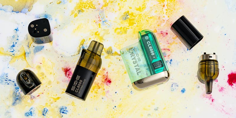 Zerlegte Komponenten einer SKE Crystal CL6000 Hybrid-Vape liegen auf einem abstrakten, bunten Hintergrund mit gelben, blauen und roten Farbspritzern. Die Einzelteile umfassen den Akku, das Mundst&uuml;ck, die Kartusche und eine durchsichtige gr&uuml;ne H&uuml;lle mit dem Branding SKE Crystal CL6000. Die Teile sind in einer schwebenden Anordnung dargestellt.