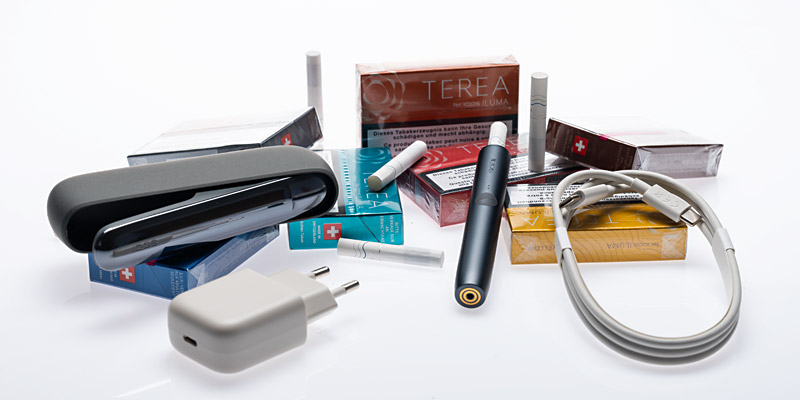 Ein Iqos-Iluma-Set bestehend aus einem schwarzen Iqos-Iluma-Ger&auml;t, einem passenden schwarzen Etui, einem wei&szlig;en USB-Ladekabel und einem Netzadapter. Im Hintergrund sind mehrere bunte Packungen Terea-Tabaksticks f&uuml;r Iqos Iluma in verschiedenen Geschmacksrichtungen zu sehen. Einige Tabaksticks liegen lose vor den Packungen. Die Packungen tragen Gesundheitswarnhinweise und das Schweizer Kreuz-Symbol. Alles liegt auf einer weissen, gl&auml;nzenden Oberfl&auml;che.