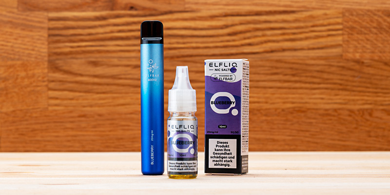Eine ELFLIQ Nic Salts E-Liquid-Flasche in der Geschmacksrichtung Blueberry steht neben einer grösseren ELFBAR Einweg-E-Zigarette in der gleichen Geschmacksrichtung. Beide Produkte sind in ihren Verpackungen zu sehen.