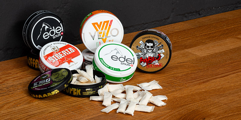 Verschiedene Snus-Dosen von Marken wie Edel, Pablo, Velo, Superwhite und Scoper auf einem Holztisch, mit ge&ouml;ffneten Dosen und sichtbaren Nikotinbeuteln.