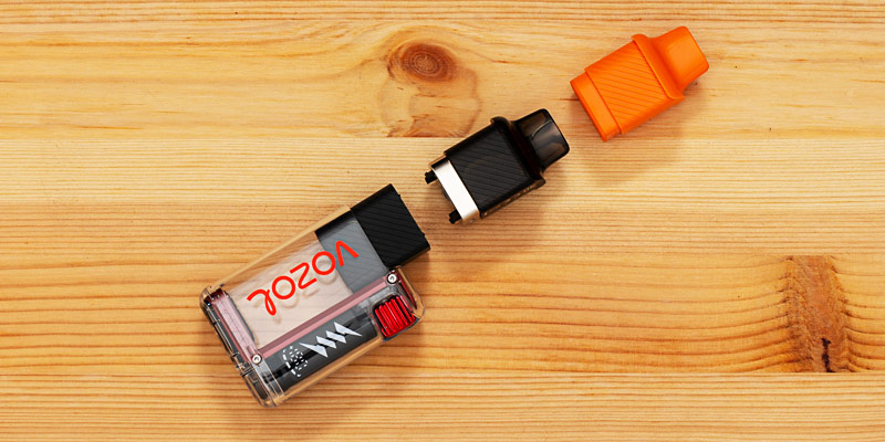 Ein auf Holz liegendes Einweg-Vape-Gerät, der Vozol Vista Plug, bestehend aus mehreren Teilen: ein durchsichtiger Hauptkörper mit der Aufschrift „Vozol“, ein schwarzer Mundstückaufsatz, ein herausnehmbarer schwarzer Anschluss, sowie eine orangefarbene Schutzkappe, die daneben liegt.