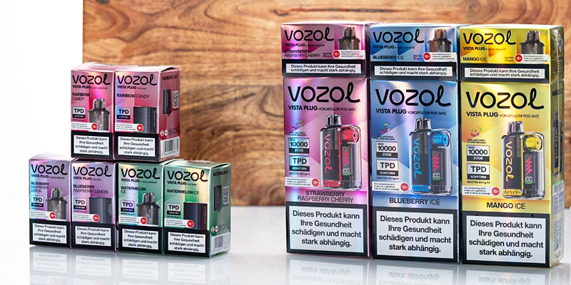 Ein Sortiment von Vozol-Vape-Produkten der Serie Vista Plug, die in bunten Verpackungen auf einer reflektierenden Oberfläche präsentiert werden. Links kleinere Schachteln mit Geschmacksrichtungen wie «Rainbow Candy» und «Watermelon Ice». Rechts grössere Verpackungen mit Geschmacksrichtungen wie «Strawberry Raspberry Cherry», «Blueberry Ice» und «Mango Ice». Auf den Verpackungen befindet sich ein Warnhinweis: «Dieses Produkt kann Ihre Gesundheit schädigen und macht stark abhängig.» Im Hintergrund eine Holzstruktur.
