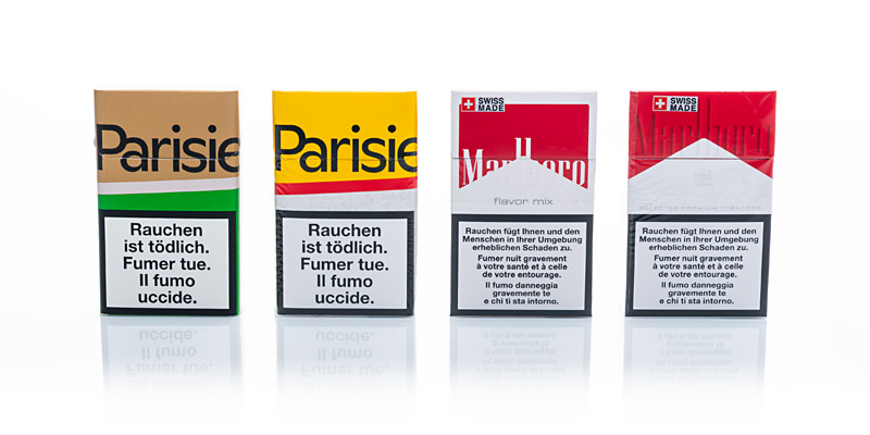 Vier Zigarettenpackungen der Marken Parisienne und Marlboro stehen in einer Reihe auf weissem Hintergrund. Auf jeder Packung ist ein großer Warnhinweis in Deutsch, Französisch und Italienisch sichtbar, der vor den Gefahren des Rauchens warnt.