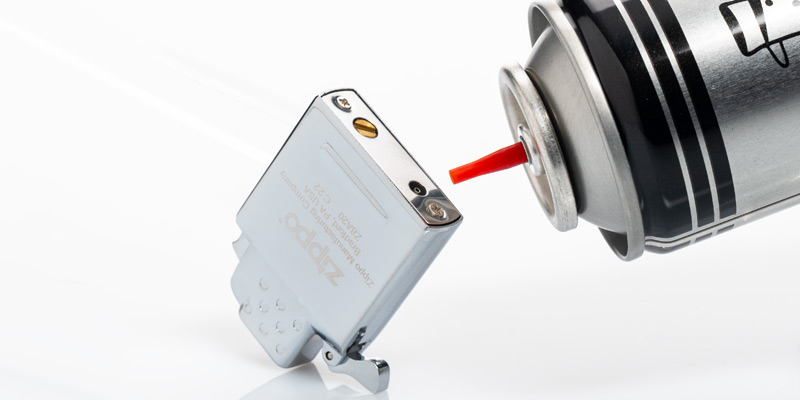 Eine Butan-Gas-Flasche, die mit Ventil schräg an die Gas-Zufuhr des Zippo-Feuerzeugs gehalten wird, um zu zeigen, dass es auch Zippos mit Gas-Inlays gibt.