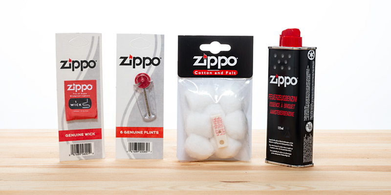 Vier Zippo-Zubeh&ouml;r-Artikel, die aufrecht auf einem Holztisch stehen: links eine Zippo-Z&uuml;ndschnur, gefolgt von einem Zippo-Feuerstein, Zippo-Watte und Zippo-Benzin (von links nach rechts).