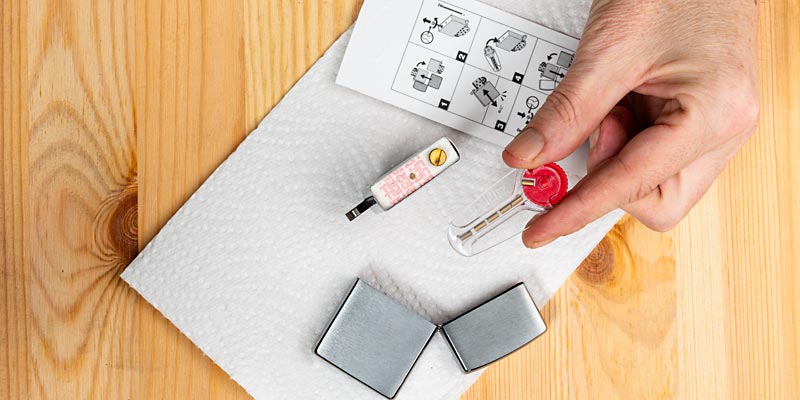 Eine Hand wechselt den Feuerstein eines Zippo-Feuerzeugs. Auf einem Papiertuch liegen die Feuerzeughülle, der Einsatz, ein neuer Feuerstein in einem transparenten Behälter und eine Bedienungsanleitung mit visuellen Schritten. Der Fokus liegt auf der präzisen Arbeit, die den Wartungsprozess zeigt. Die Szene spielt sich auf einer Holzoberfläche ab.
