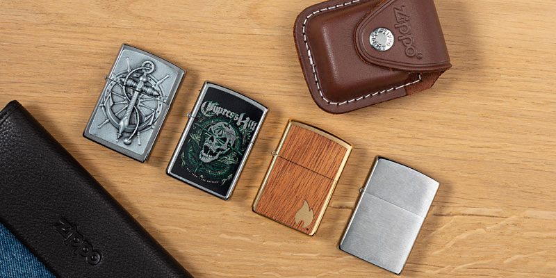 Vier aneinandergereihte Zippo-Feuerzeuge auf einem Holztisch, eines zeigt ein Cypress-Hill-Logo, eines eine Holz-Hülle, ein weiteres eine metallene Aussenhülle, während die metallene Hülle des vierten Zippos wird von einem Seemanns-Haken bekleidet ist. Daneben liegt ein Zippo-Leder-Etui.