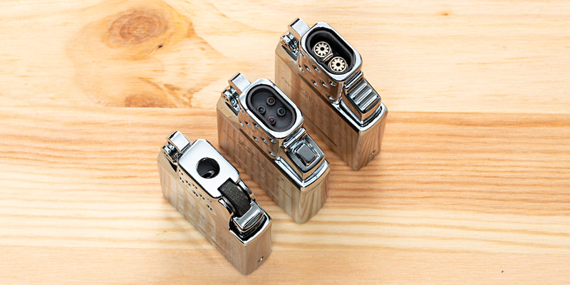 Drei verschiedene Einsätze für Zippo-Feuerzeuge auf einer hellen Holzoberfläche. Die Einsätze haben unterschiedliche Mechanismen: einer mit Feuersteinrad, einer mit Plasma-Zündung und einer mit Gasbrenner. Sie glänzen in einem polierten Metall-Finish und sind sorgfältig angeordnet, um ihre Funktionalität und Vielfalt zu zeigen.