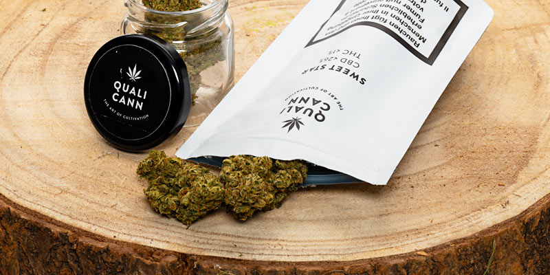 Qualicann-CBD-Blüten: Glas und Packung geöffnet auf Holzstrunk.