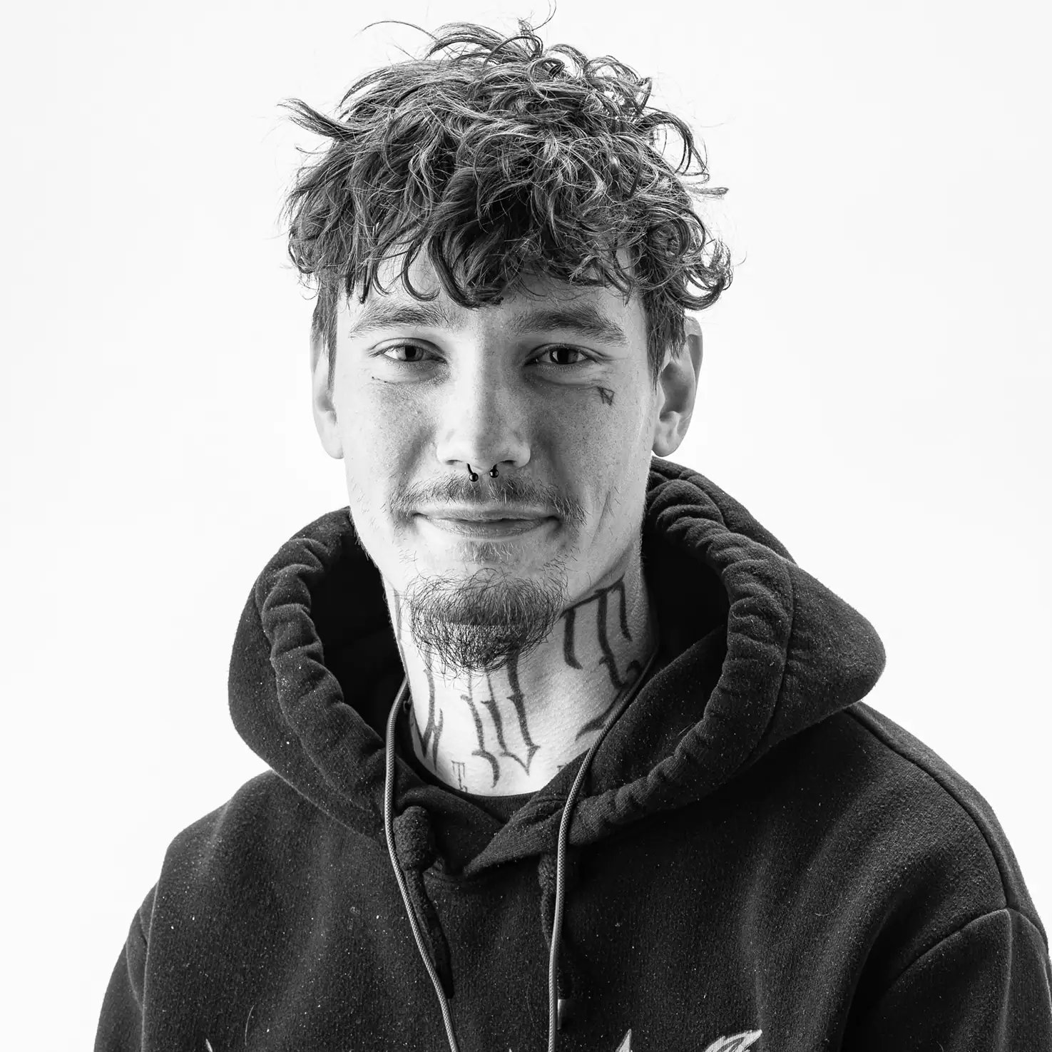 Porträt eines jungen Mannes mit lockigem Haar, Gesichtstattoos und Kapuzenpulli, vor hellem Hintergrund.
