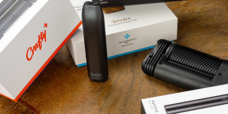 Verschiedene Vaporizer-Modelle auf einem Holztisch, darunter Geräte von iven, Wolkenkraft, Storz & Bickel und Puffco, jeweils mit ihren Verpackungen im Hintergrund.