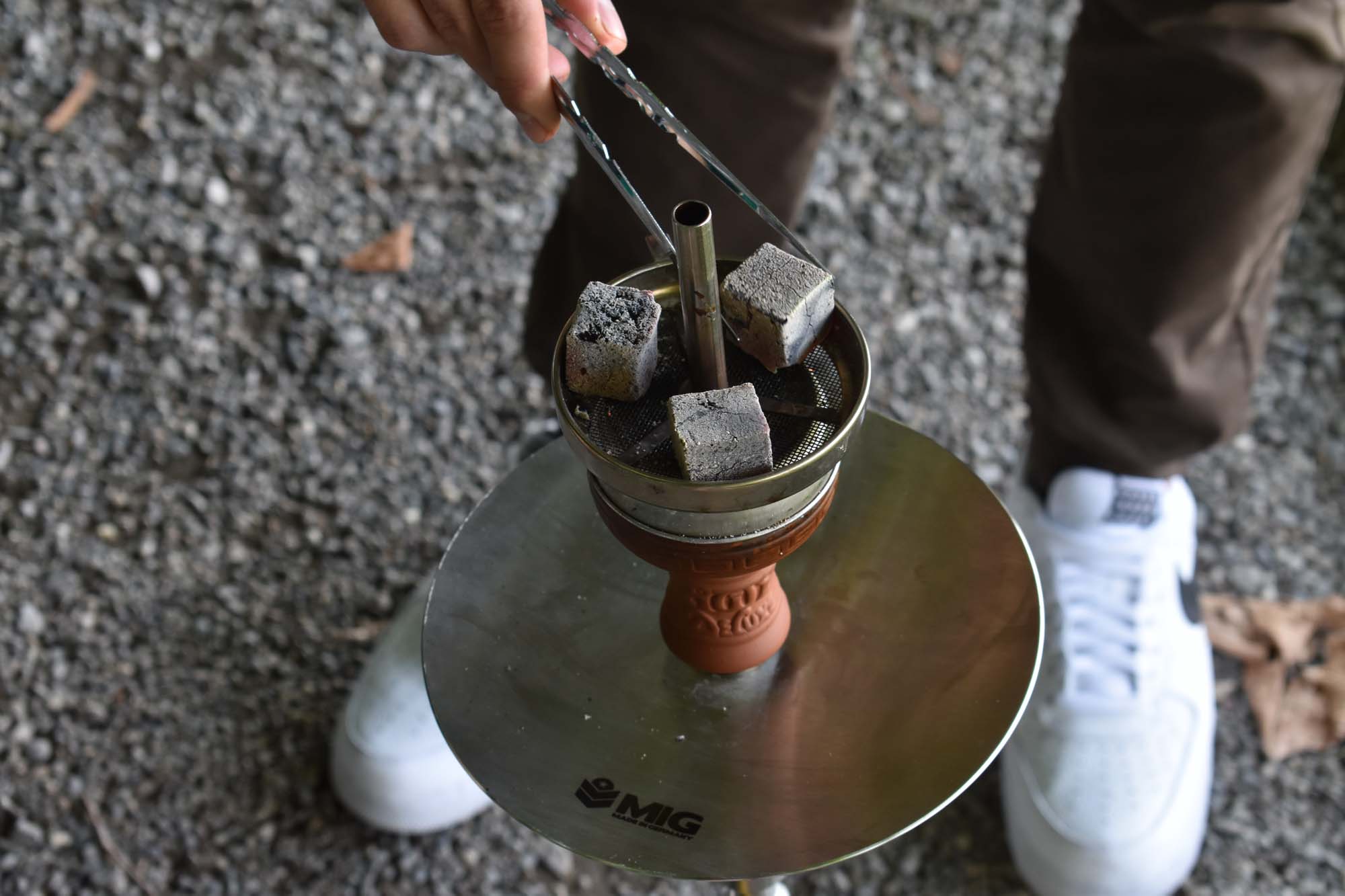 Hand platziert heisse Naturkohle mit Zange auf Shisha-Kopf mit Kaminaufsatz, Kohleteller mit MIG-Logo im Freien