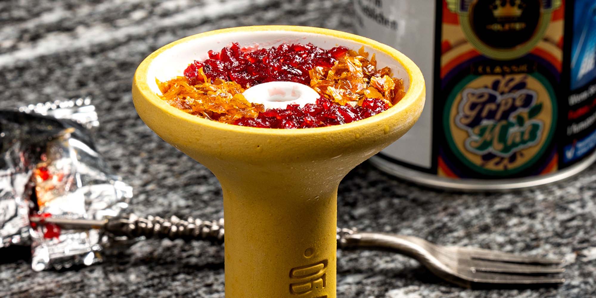 Gelber Shisha-Phunnel mit zweifarbig gemischtem Tabak. auf Granitplatte, Gabel und Tabakdose im Hintergrund