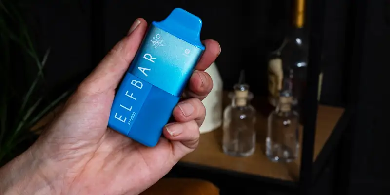 Hand h&auml;lt einen blaue Elfbar AF5000 Hybrid-Vape im Vordergrund. Im Hintergrund stehen Glasflaschen und eine dunkle Einrichtung.