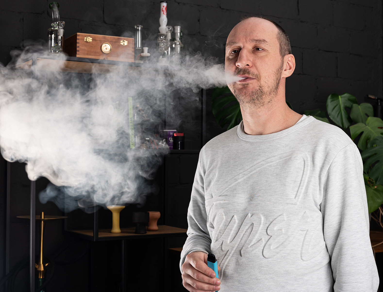 Ein Mann mit einem Vape in der Hand, der Dampf ausbl&auml;st. Im Hintergrund sind verschiedene Vaping-Ger&auml;te und Zubeh&ouml;rteile zu sehen.